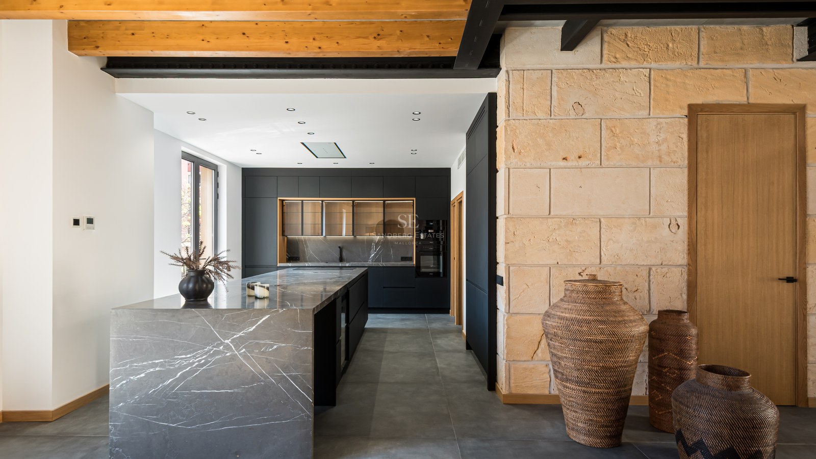 A contemporary kitchen featuring a grey marble island, black cabinetry, exposed wooden ceiling beams, and a stone wall.
