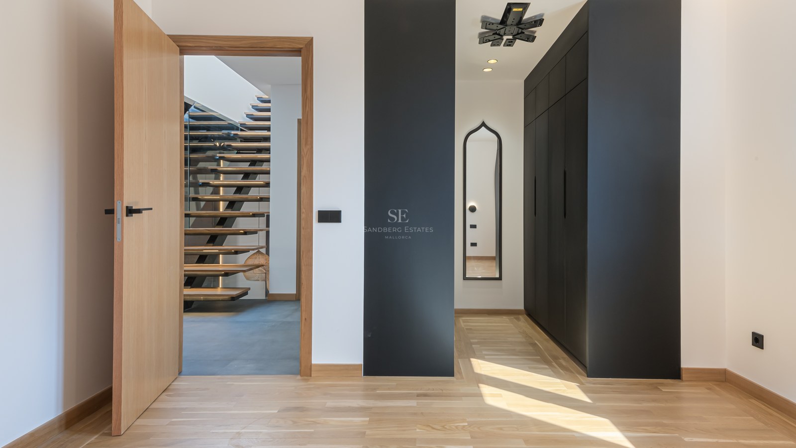 Modern hallway with light wood floors, black built-in closets, and an open door viewing a floating wooden staircase.