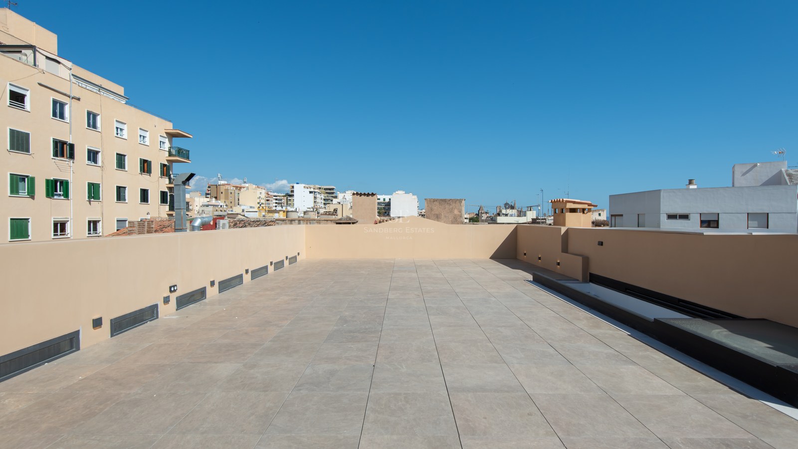 Spacious rooftop terrace with beige tiles and modern wall lighting under a clear blue sky.