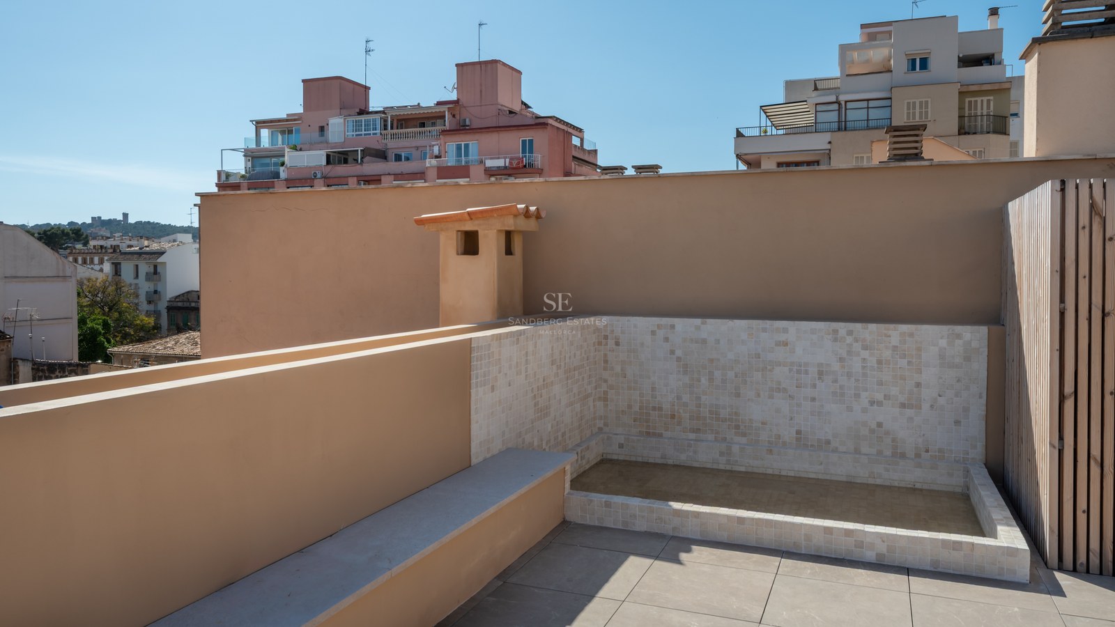 A modern rooftop terrace featuring a tiled plunge pool, beige stone flooring, and views of city buildings under a blue sky.