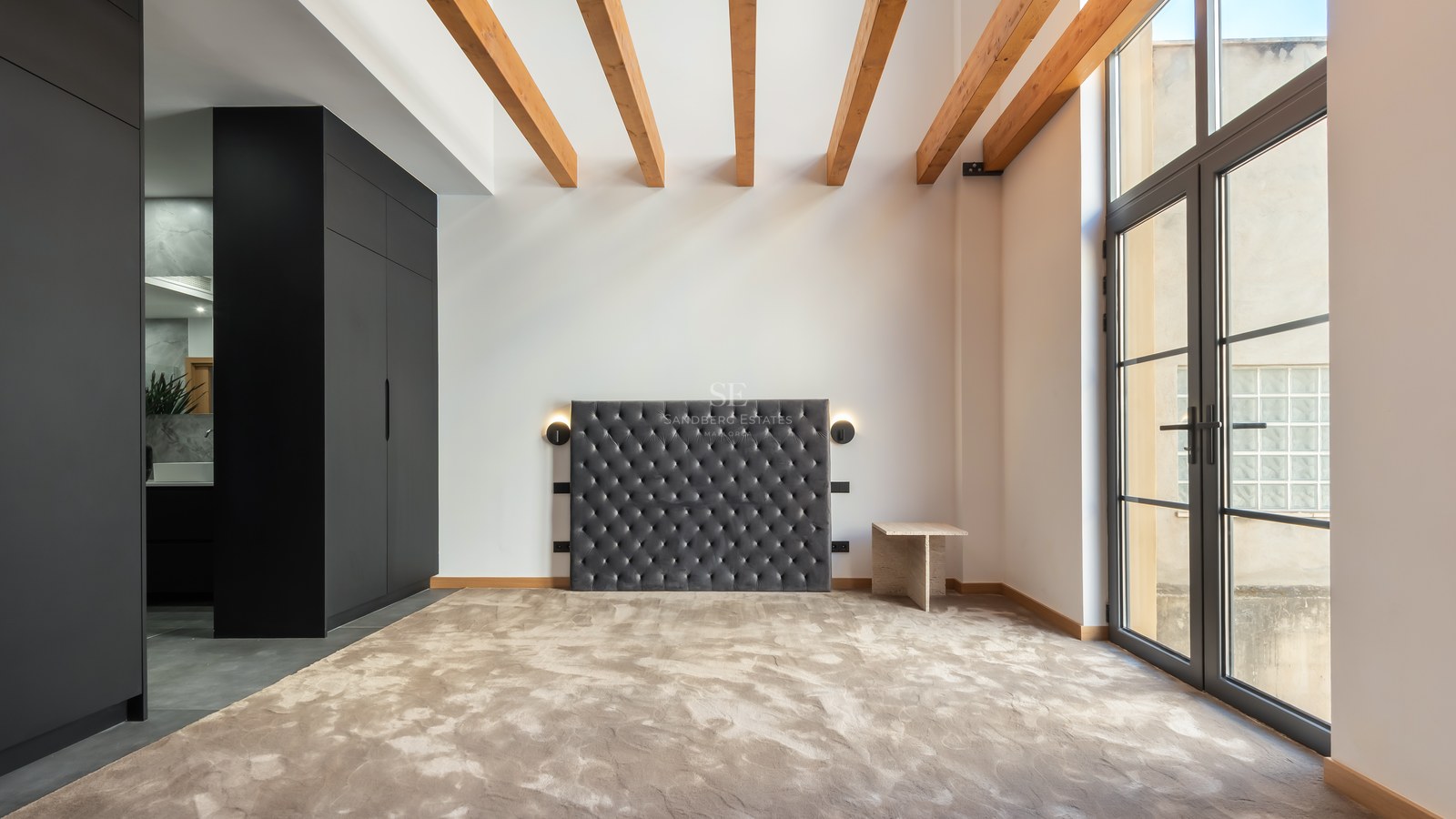 Bedroom featuring a grey velvet tufted headboard, wooden ceiling beams, black wardrobe, and large glass doors.
