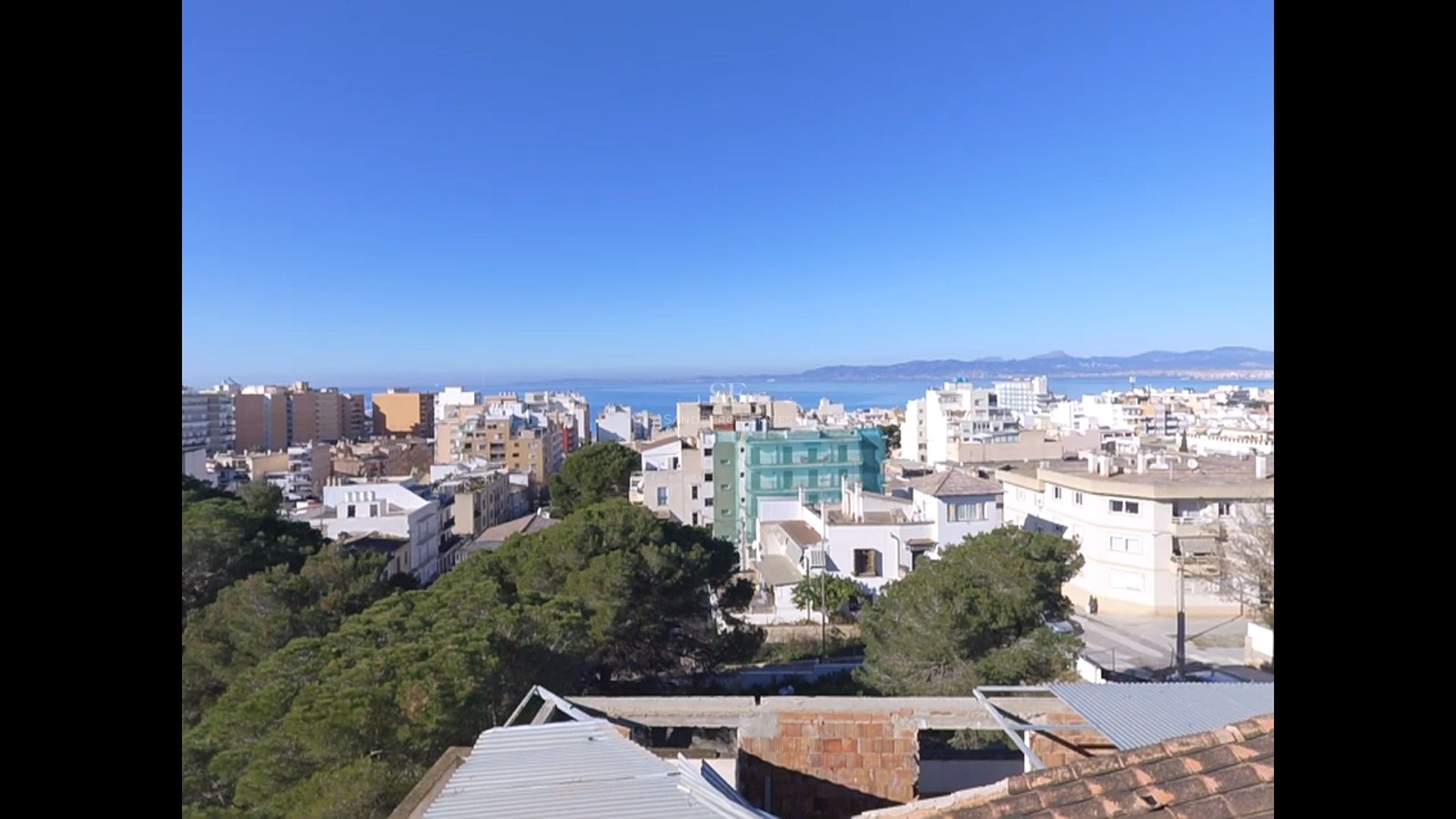 High-angle view of a coastal city with white buildings, green pine trees, and the blue Mediterranean sea in the background.