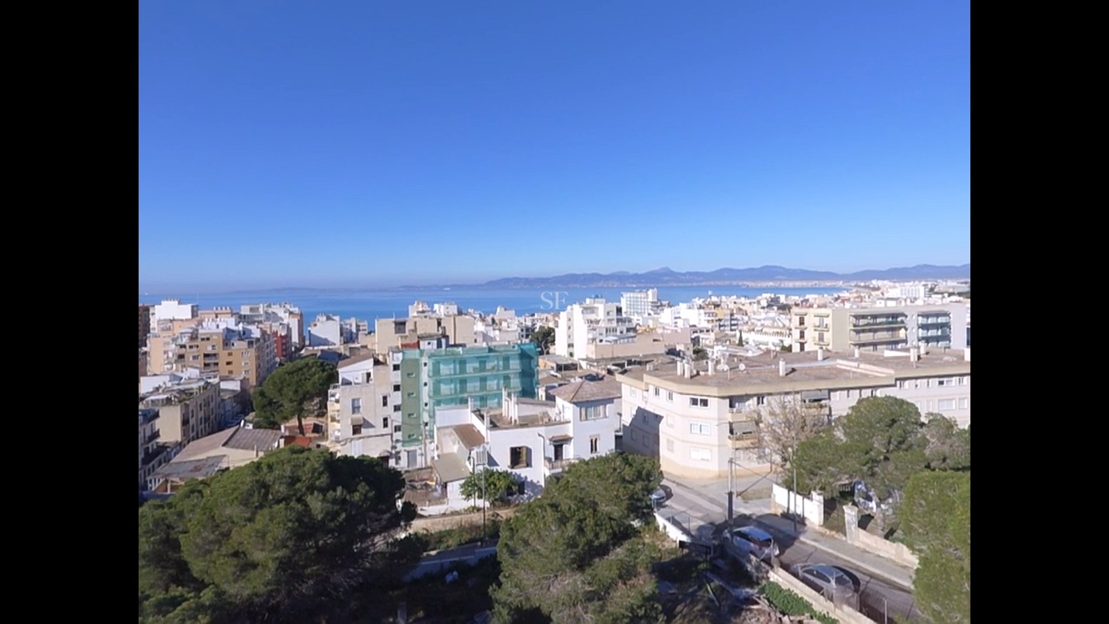 Elevated view of a Mediterranean coastal city with white buildings, green trees, and the blue sea on the horizon.