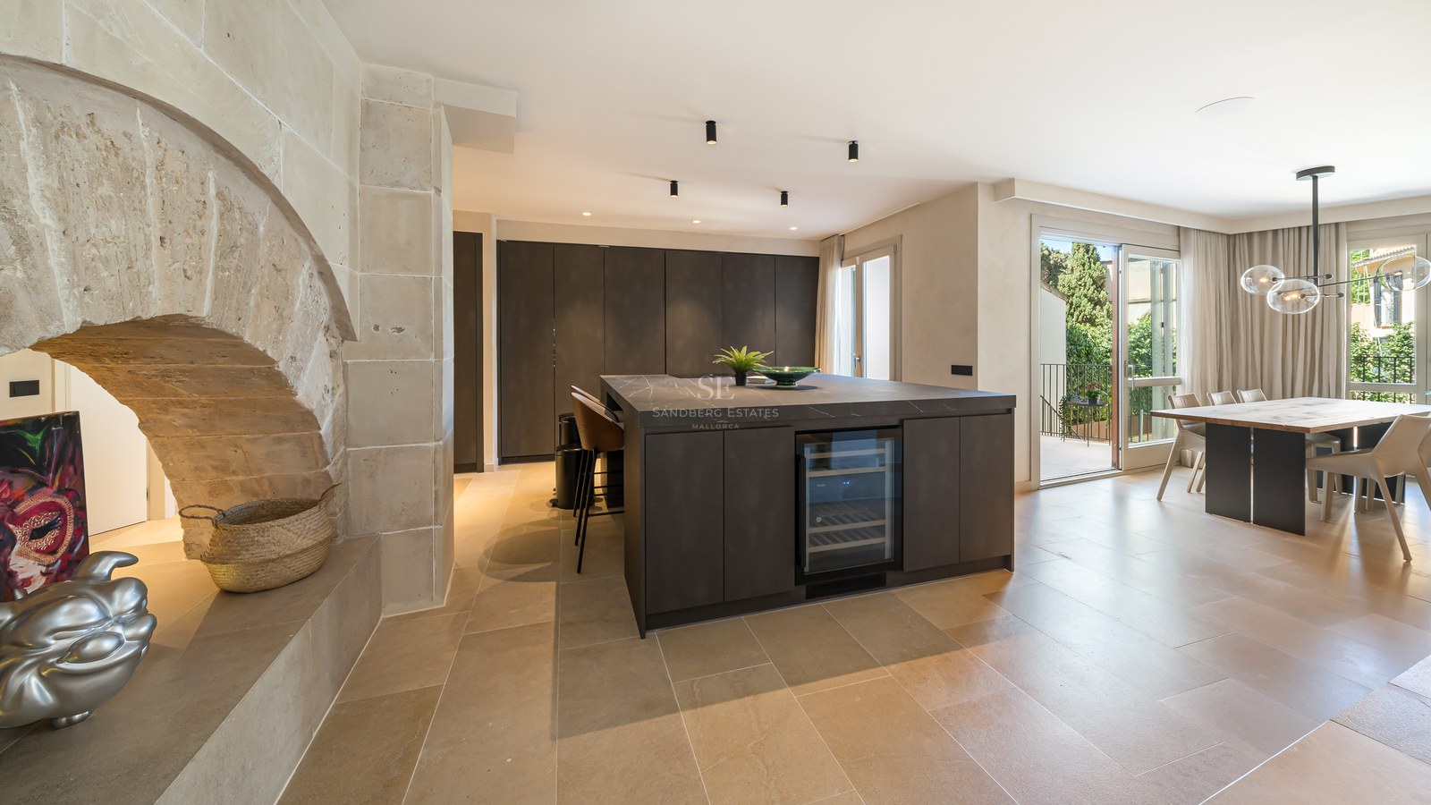Modern kitchen with dark island and wine fridge next to a rustic stone arch and open-plan dining area.