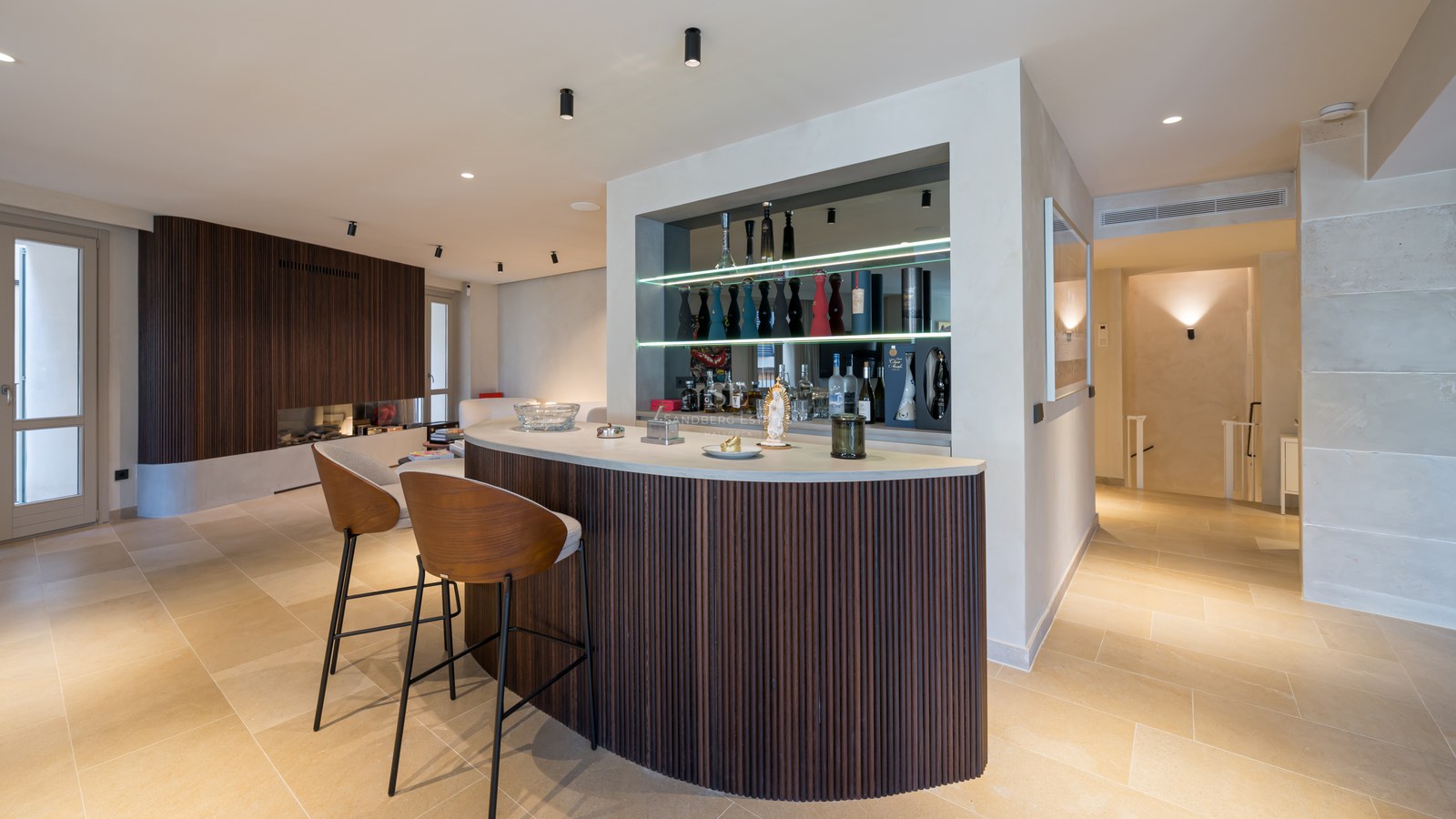 Curved home bar with dark wood slats, two designer stools, and backlit shelves with premium liquor bottles.