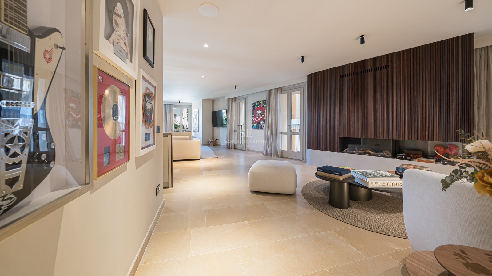 Modern living room featuring a wood-slat fireplace wall, stone floors, and framed music memorabilia in a hallway.