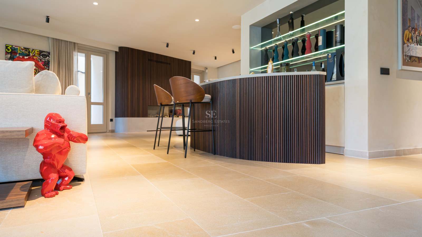 Modern living room featuring a curved wooden bar, designer stools, stone floors, and a bright red gorilla sculpture.