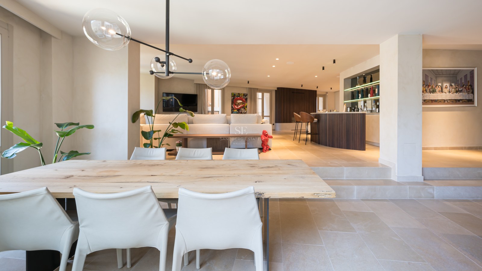 A spacious modern open-plan room featuring a wooden dining table, white chairs, glass globe pendants, and a bar area.