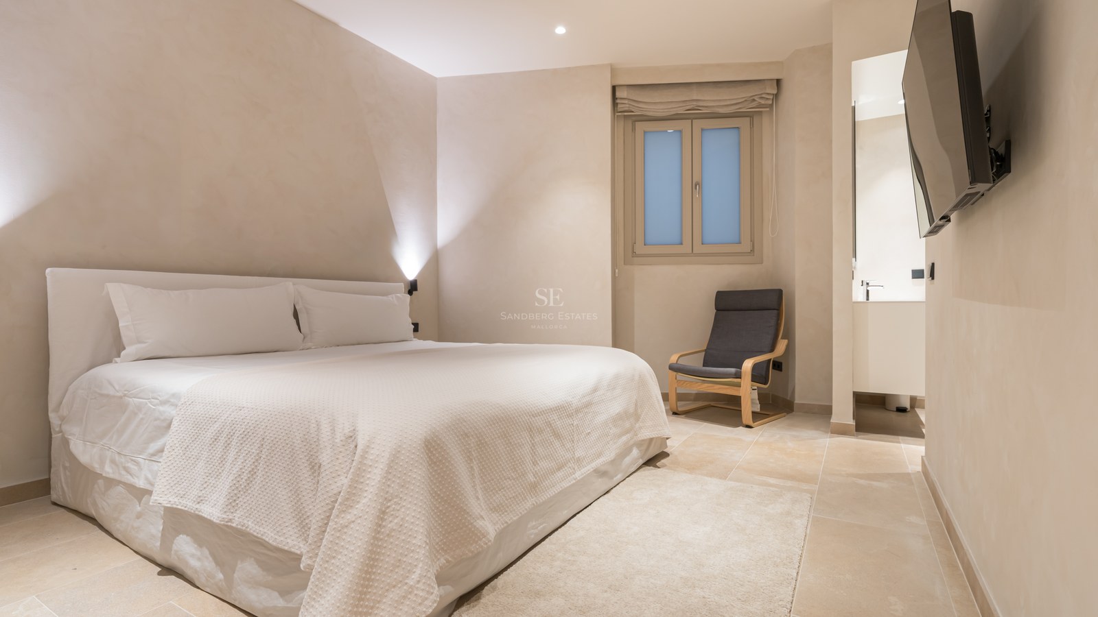 A serene bedroom with a large white bed, a grey designer chair, stone flooring, and soft beige walls.