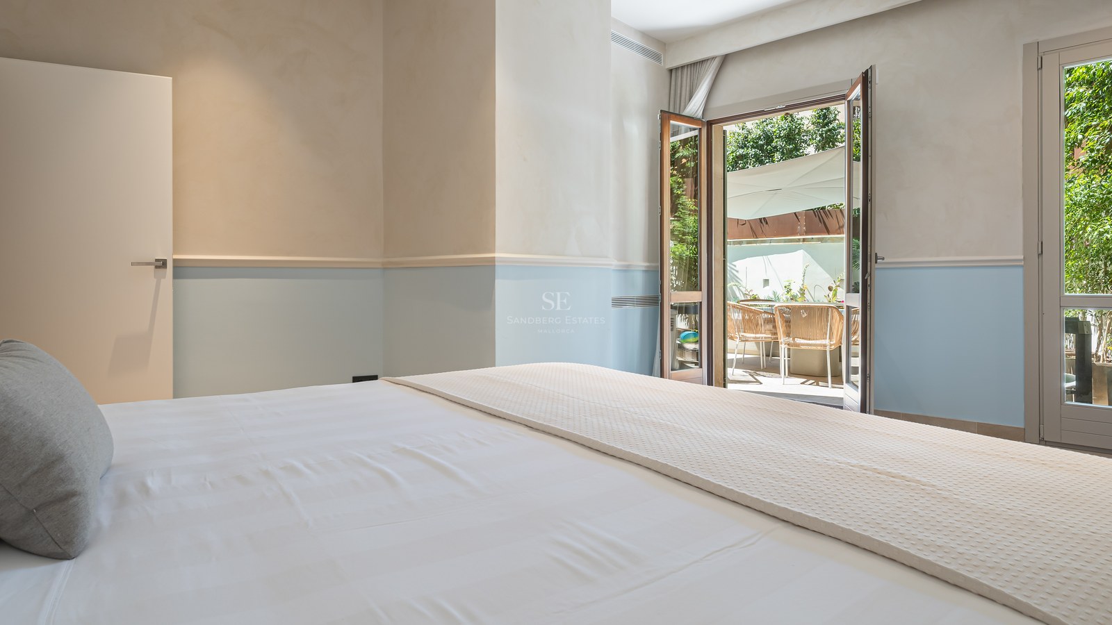 Bedroom with white linens and light blue walls looking out onto a sunny terrace through open French doors.