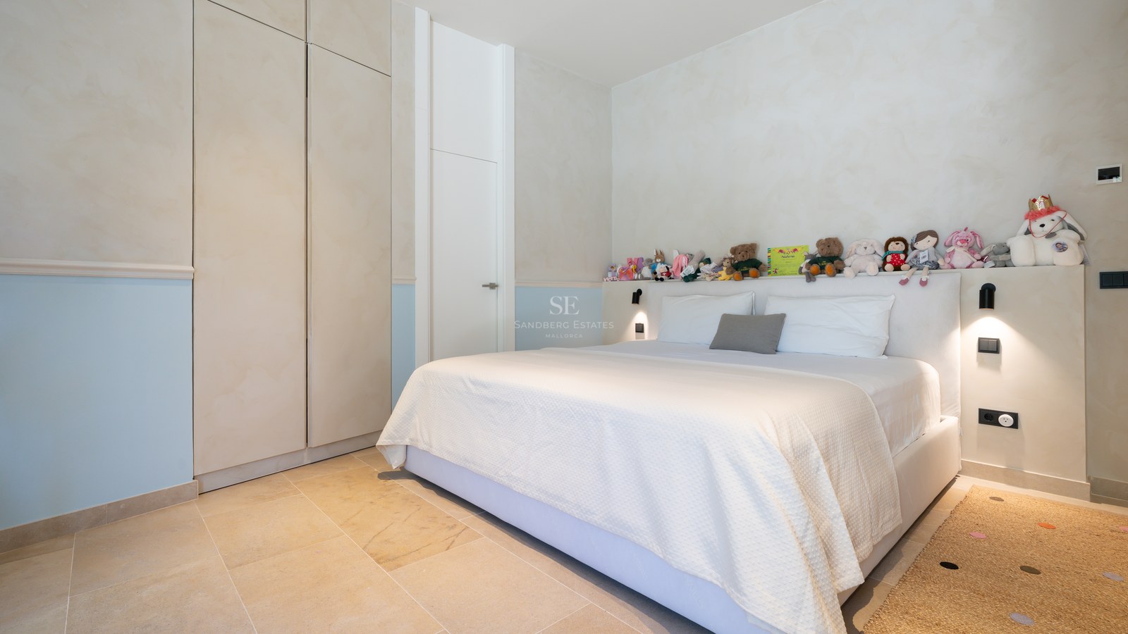 Bright bedroom featuring a large bed, stone floors, built-in wardrobes, and a row of stuffed animals on the headboard shelf.