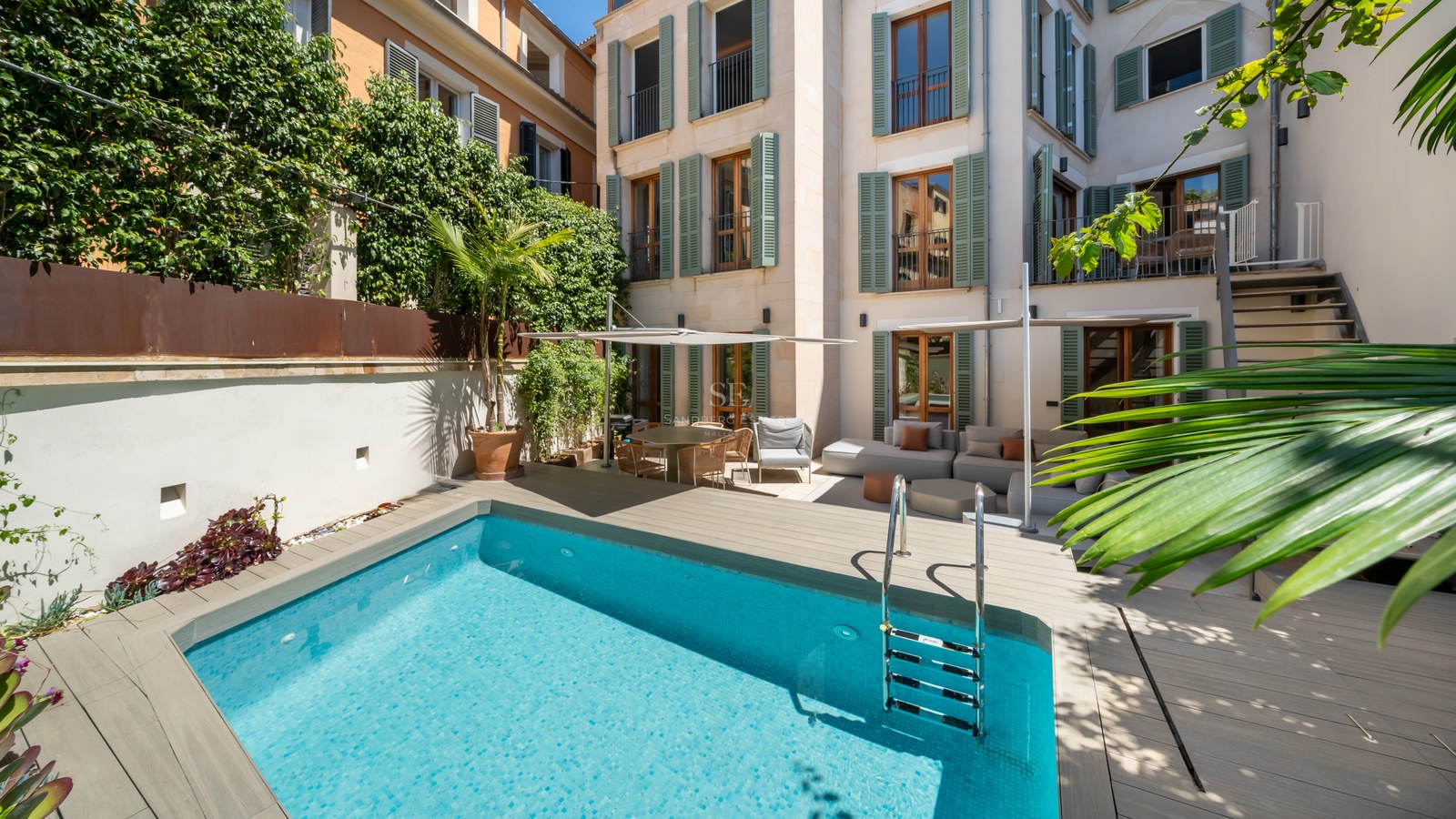 Light blue pool in a courtyard with wooden decking, outdoor sofas, and a building facade with green shutters.