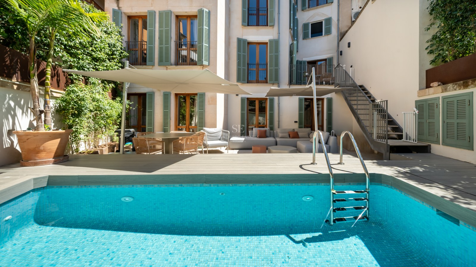 A turquoise swimming pool with a wooden deck, lounge furniture under shade sails, and a building with green shutters.
