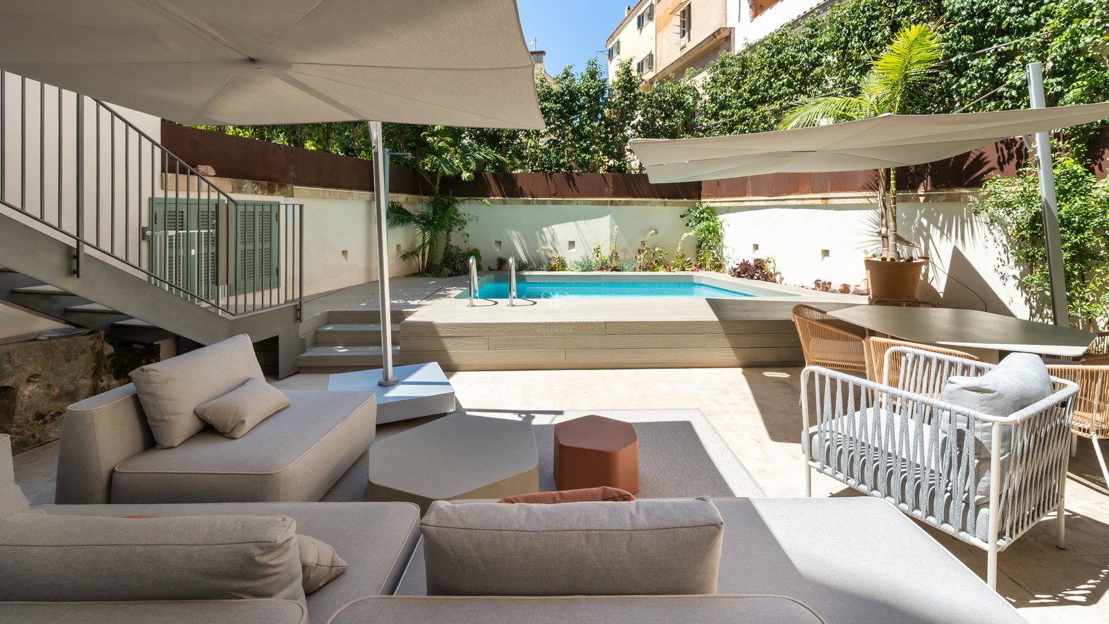 Modern terrace featuring a turquoise pool, wooden deck, grey sofas, and sun umbrellas under a clear sky.
