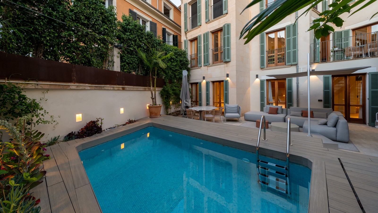 Modern private pool area with wooden decking, grey outdoor sofas, and a Mediterranean-style building with green shutters.