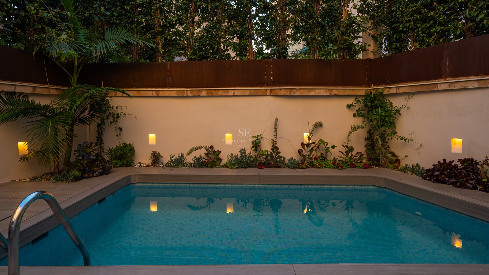 Small rectangular blue-tiled pool at dusk with warm wall lights, wood-effect decking, and lush greenery.
