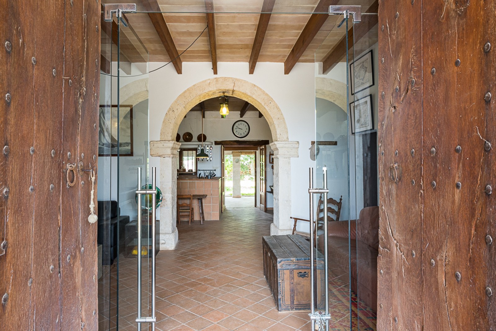 Large rustic wooden doors open to a grand foyer with a stone archway, terracotta floors, and wooden ceiling beams.