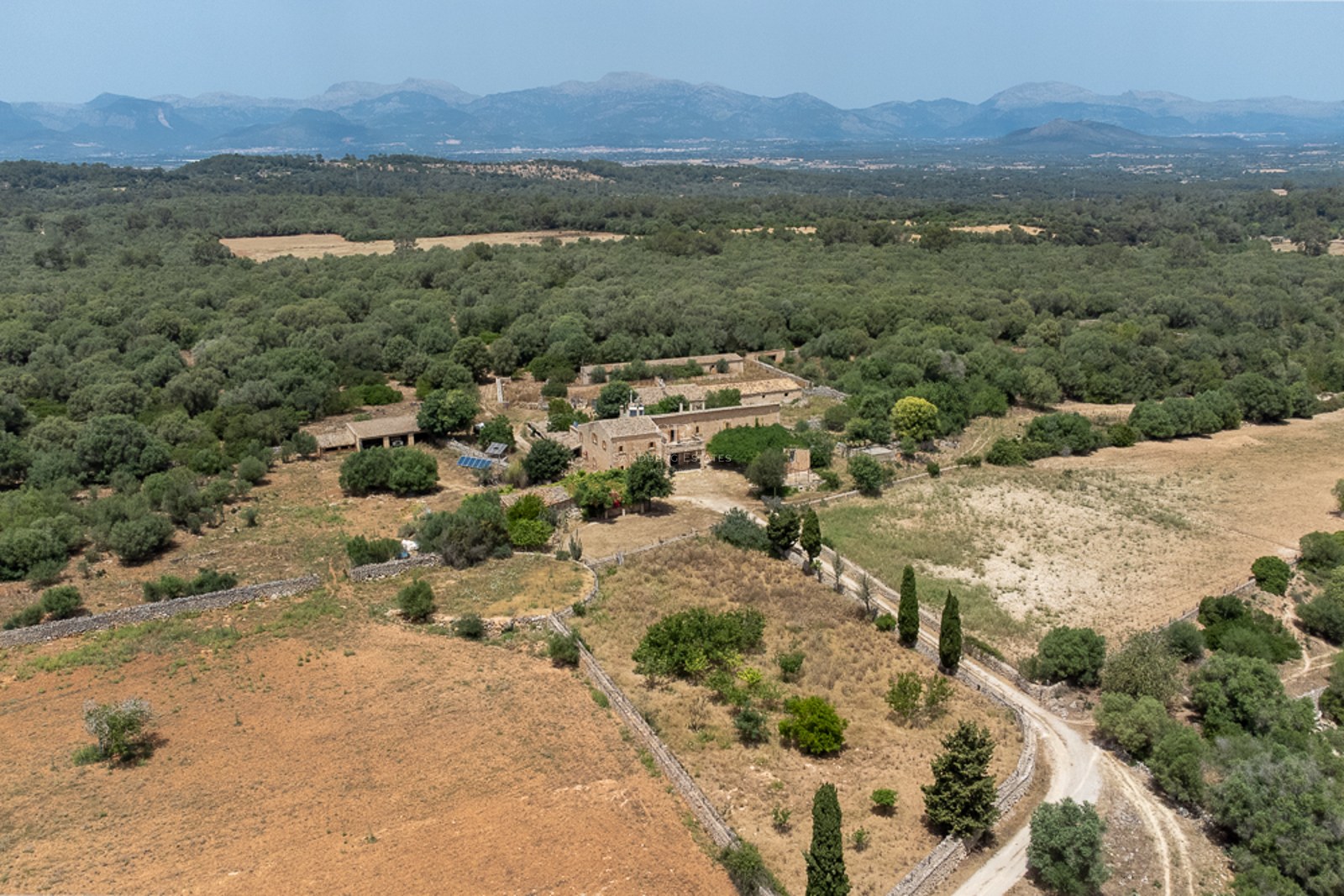 Aerial view of a large stone estate surrounded by forests and fields with mountains in the background under a clear sky.