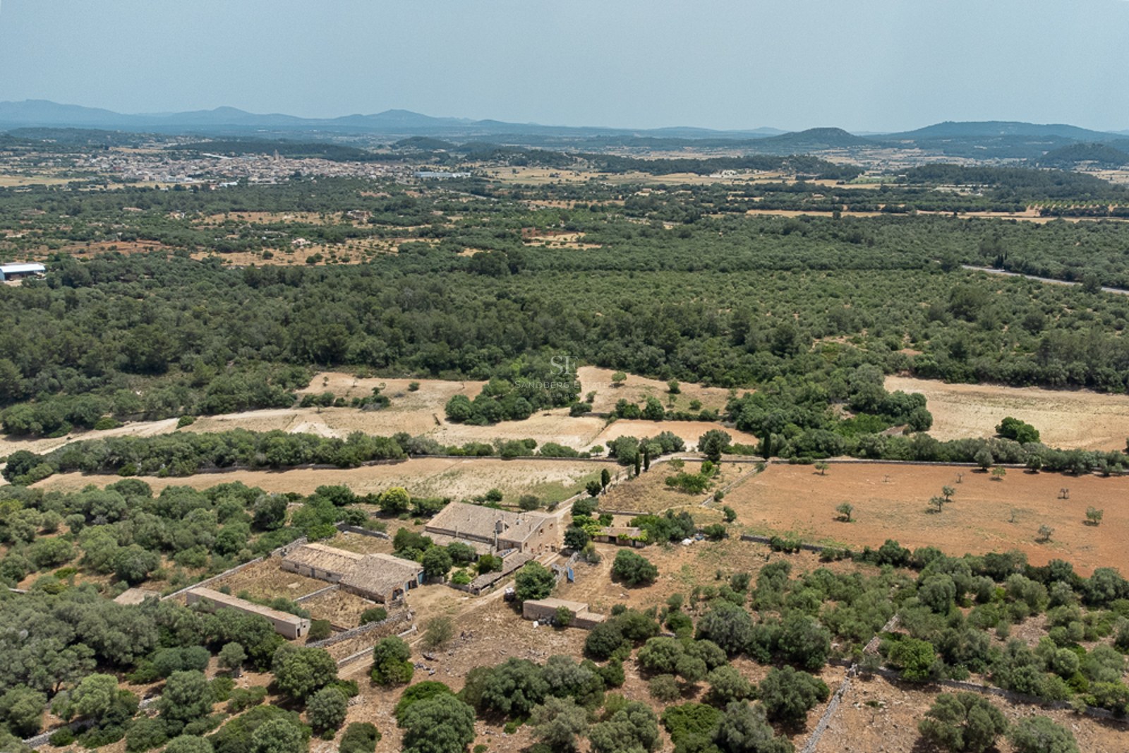 High-angle drone shot of a large stone farmhouse surrounded by lush greenery and vast fields under a clear sky.