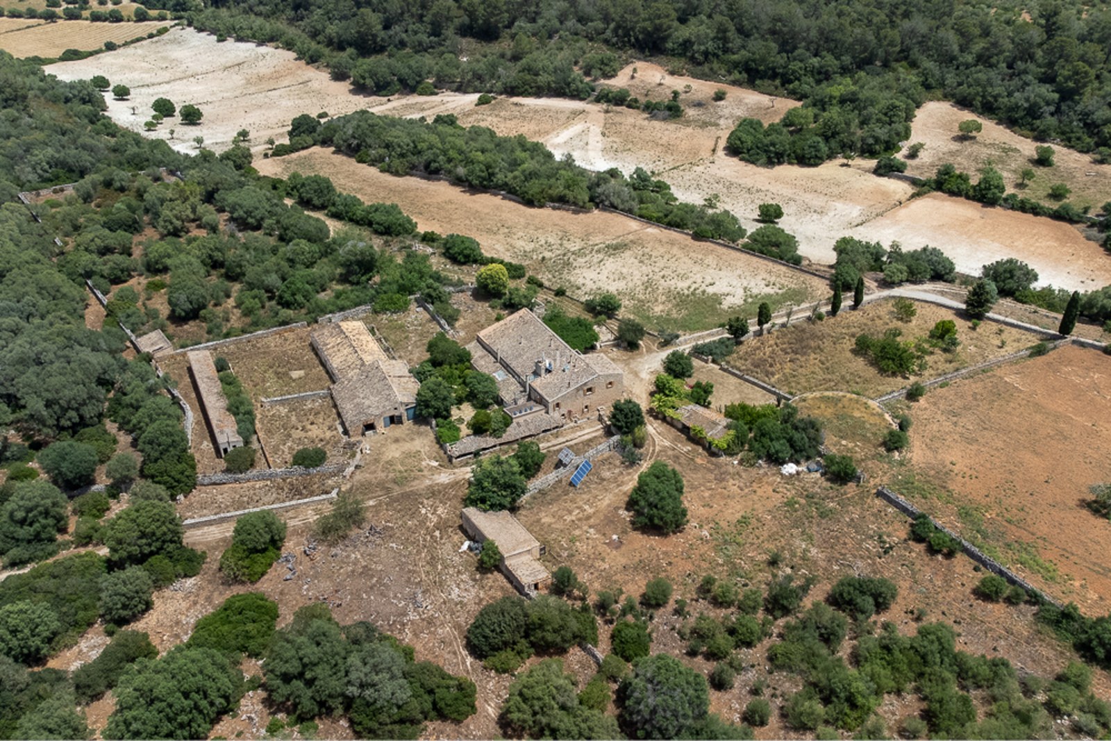 Aerial drone shot of a rustic stone farmhouse complex with multiple outbuildings surrounded by dry fields and green trees.