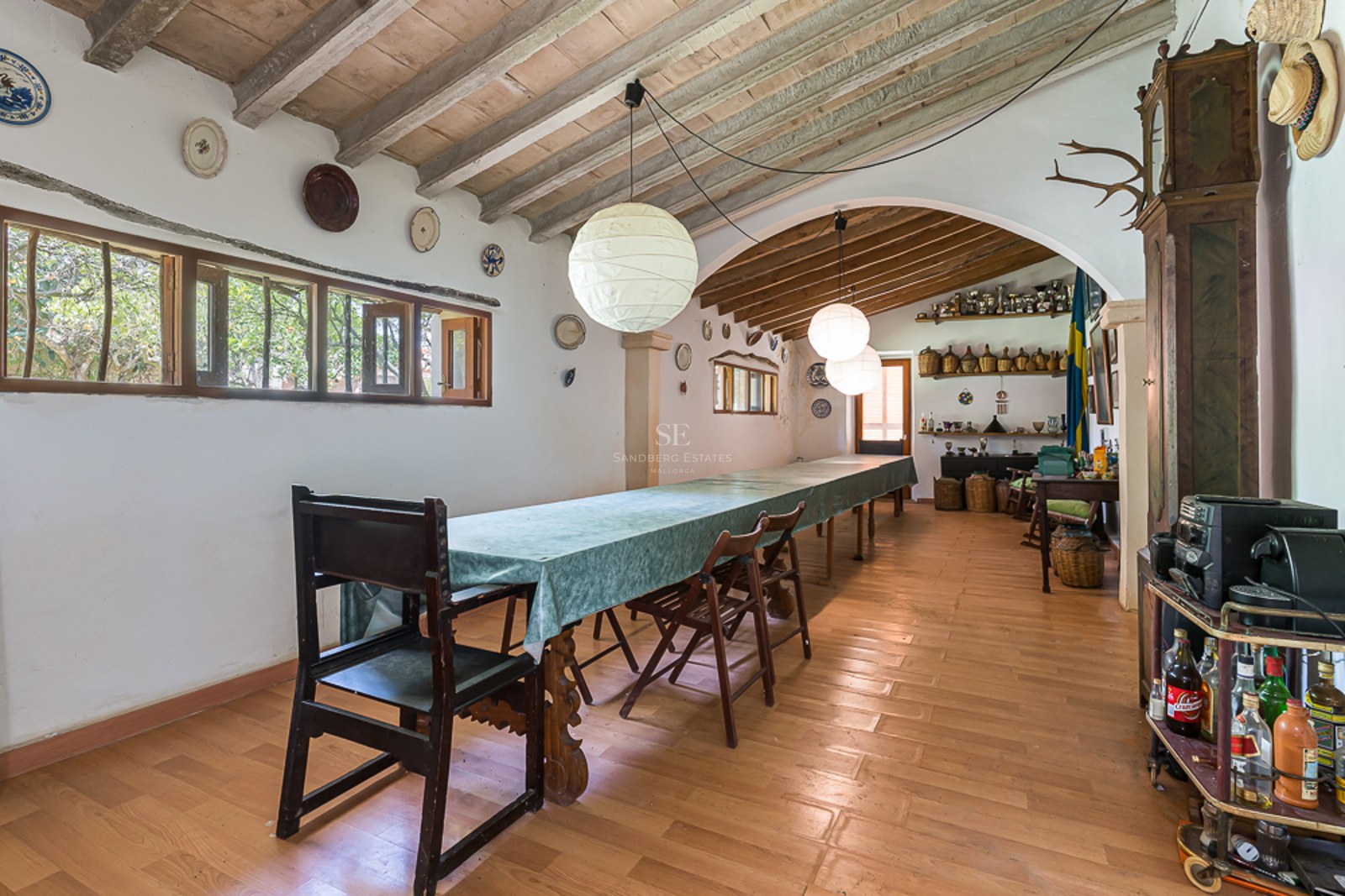 A long dining table with dark wood chairs in a rustic room with exposed beam ceilings and white plaster walls.