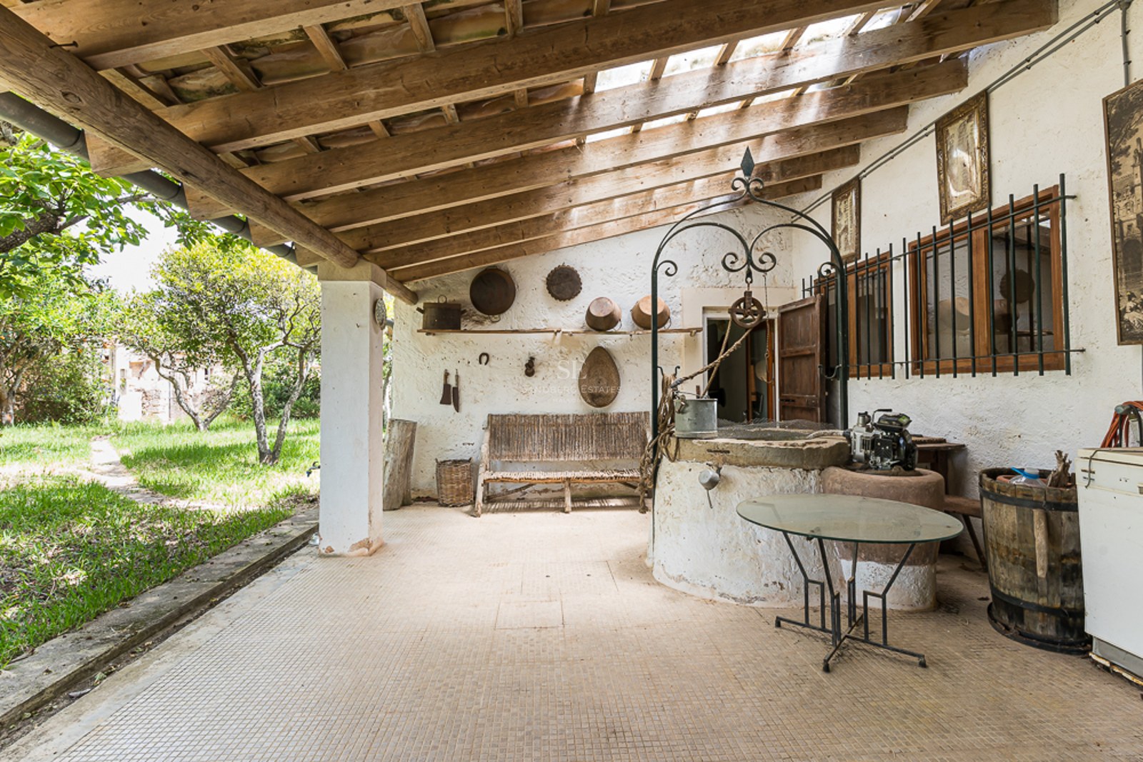 Covered terrace featuring an antique stone well, exposed wooden beams, and rustic decor overlooking a green garden.