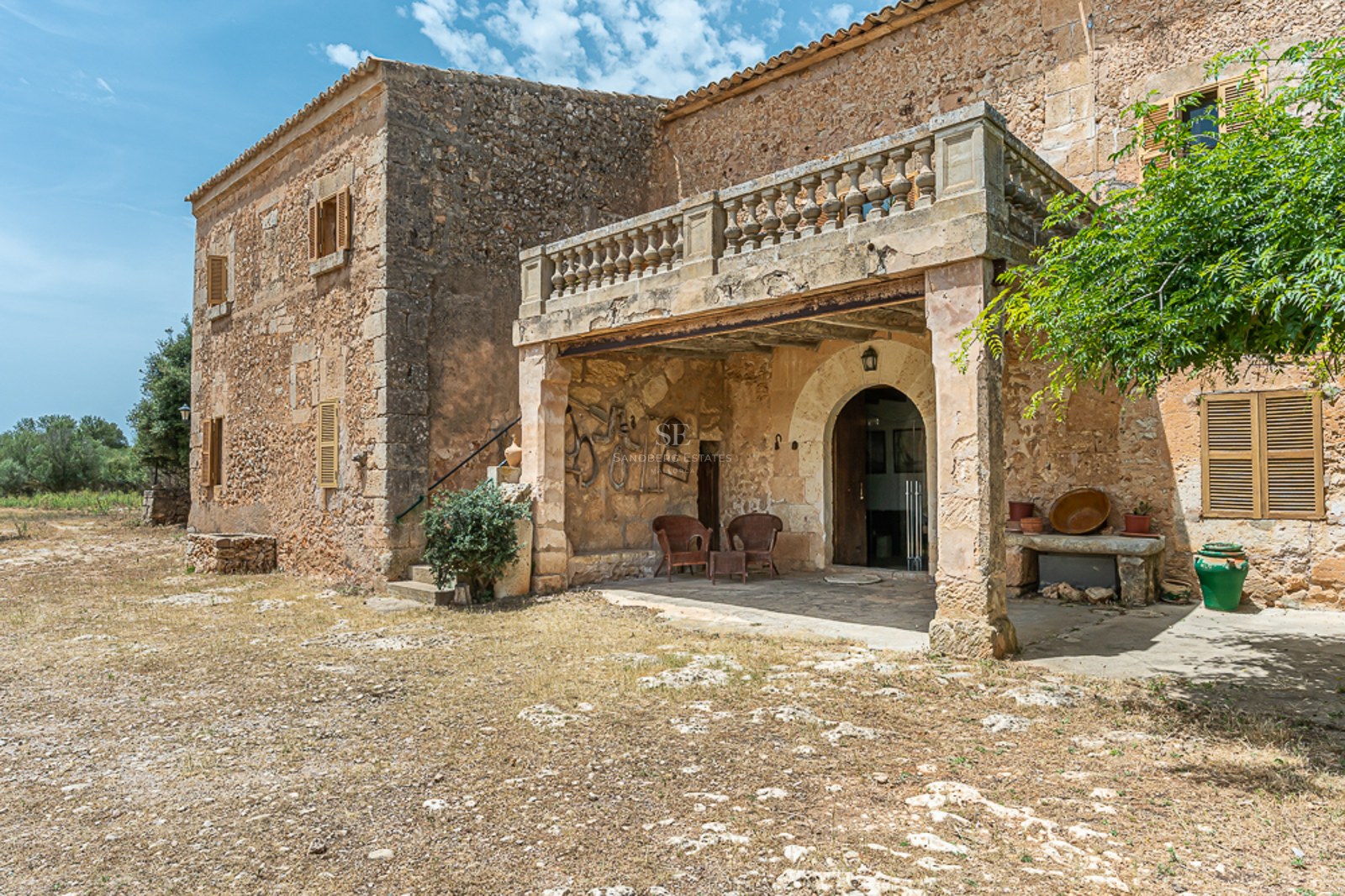 Rustic stone building with an arched entrance, stone pillars supporting a balustrade balcony under a blue sky.