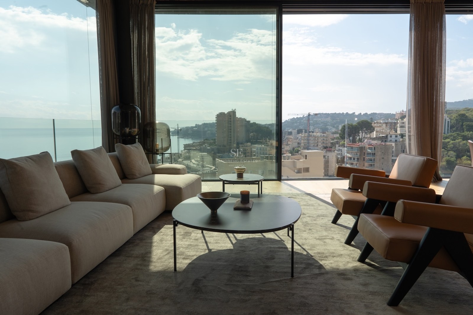 Modernes Wohnzimmer mit bodentiefen Fenstern, Blick auf die Küstenstadt, beiges Sofa und Lederstühle.