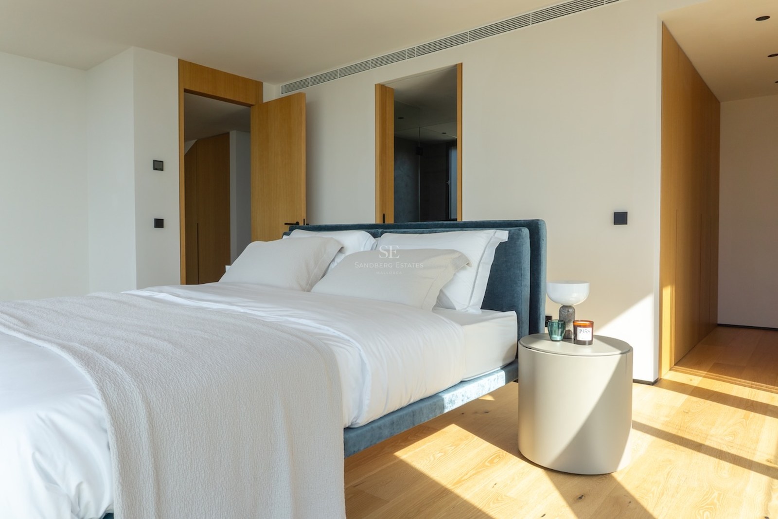 Modernes Schlafzimmer mit Eichenboden, blauem Samtbett und minimalistischen Holzelementen im Sonnenlicht.