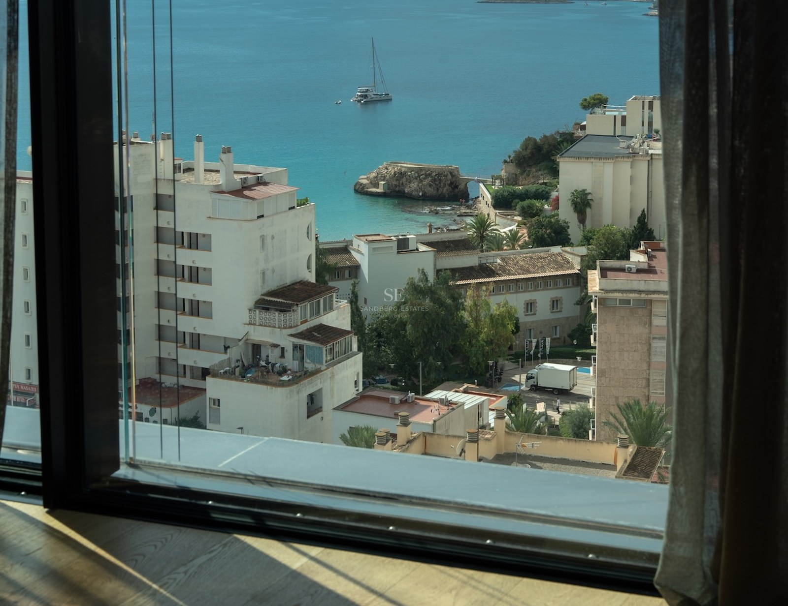 View from a modern window overlooking a turquoise bay with a sailboat and white Mediterranean buildings.