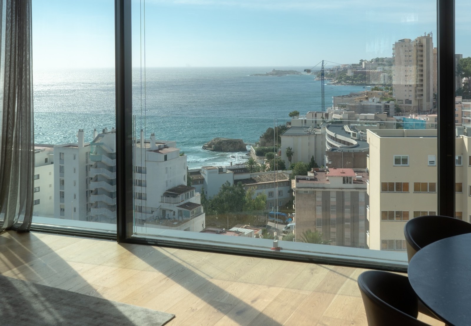 Interior view through floor-to-ceiling windows showing the Mediterranean coast, blue sea, and light wood flooring.