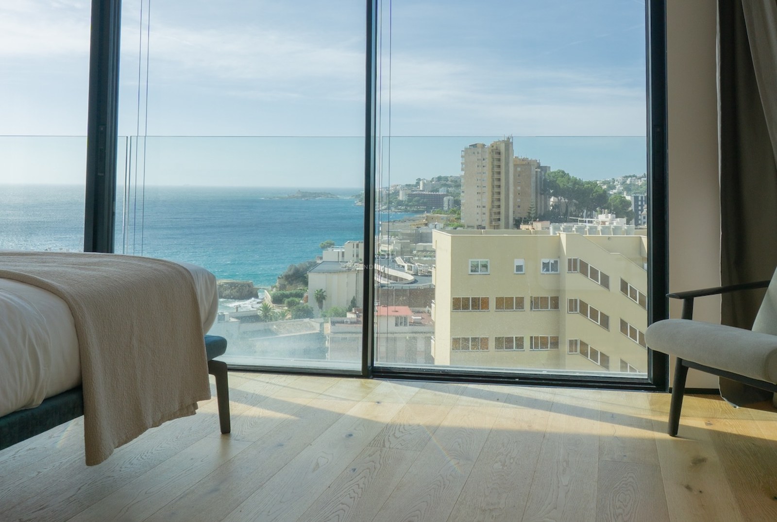 Interior of a minimalist bedroom featuring floor-to-ceiling windows overlooking the Mediterranean coast and city.