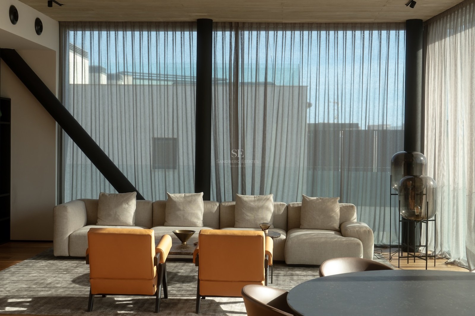 Modern living room with a large beige sofa, two tan leather armchairs, and floor-to-ceiling windows with sheer curtains.