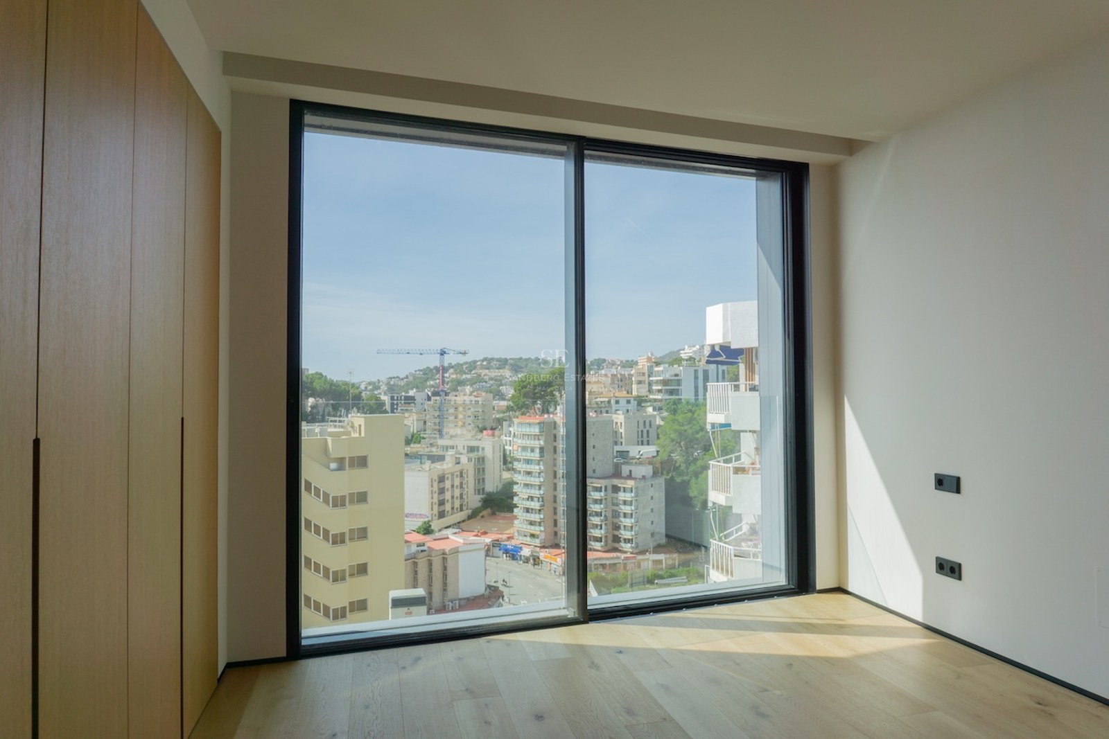 Leeres modernes Schlafzimmer mit hellen Eichenböden, Einbauschränken und einem großen bodentiefen Fenster mit Stadtblick.