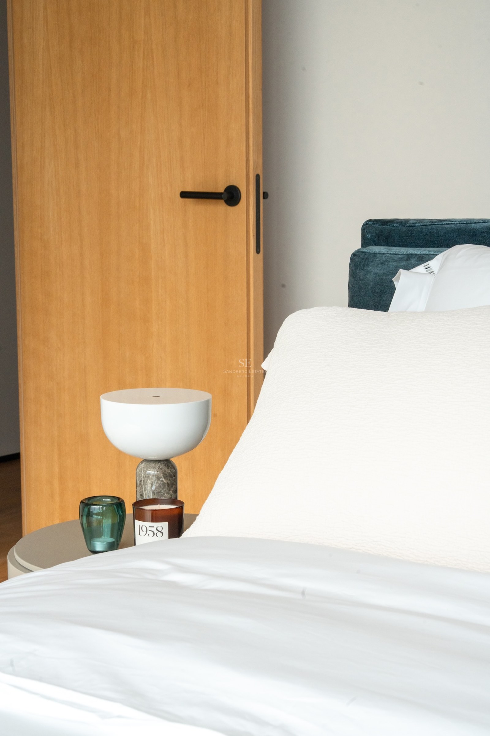 Close-up of a bed with white linens, teal velvet headboard, marble lamp, and natural oak door.