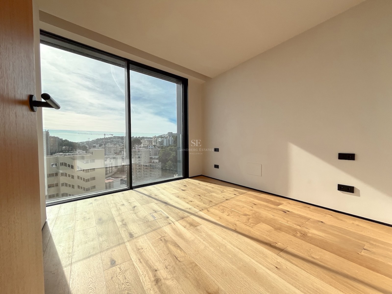 Empty modern bedroom featuring floor-to-ceiling glass doors, light oak wood floors, and panoramic city views.