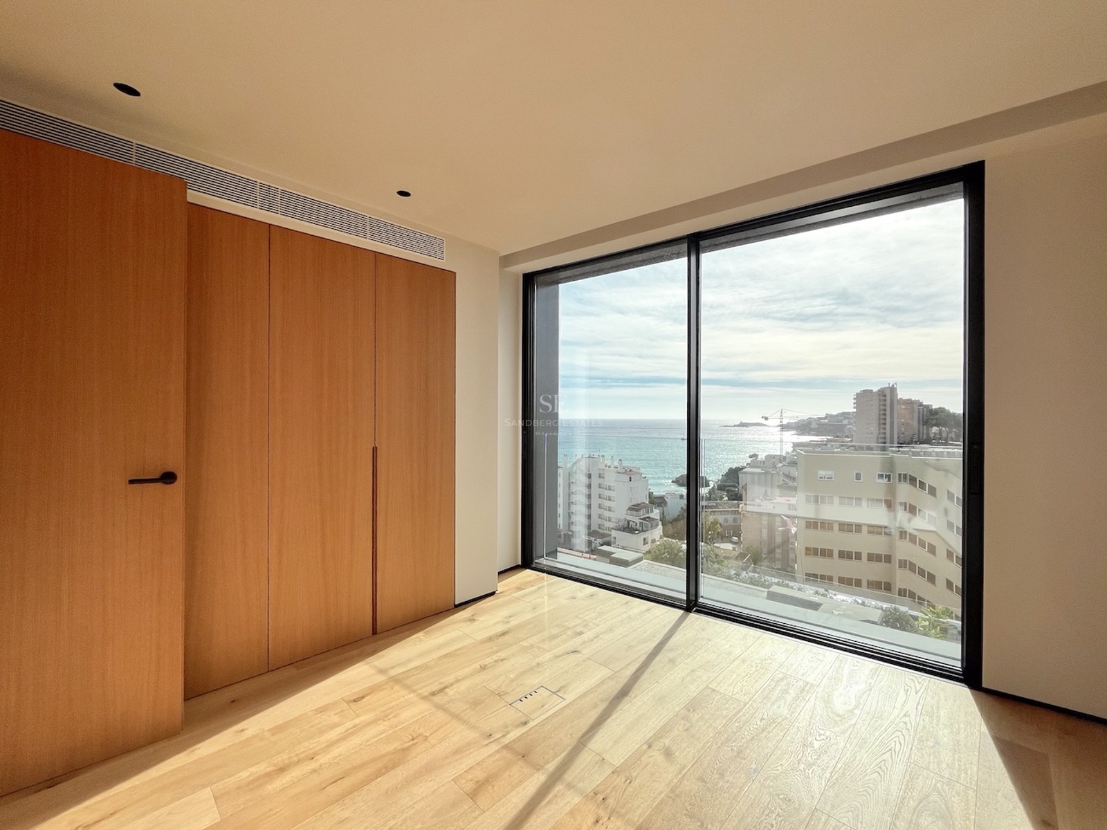 Bright empty bedroom with floor-to-ceiling windows overlooking the Mediterranean coast and built-in oak wardrobes.