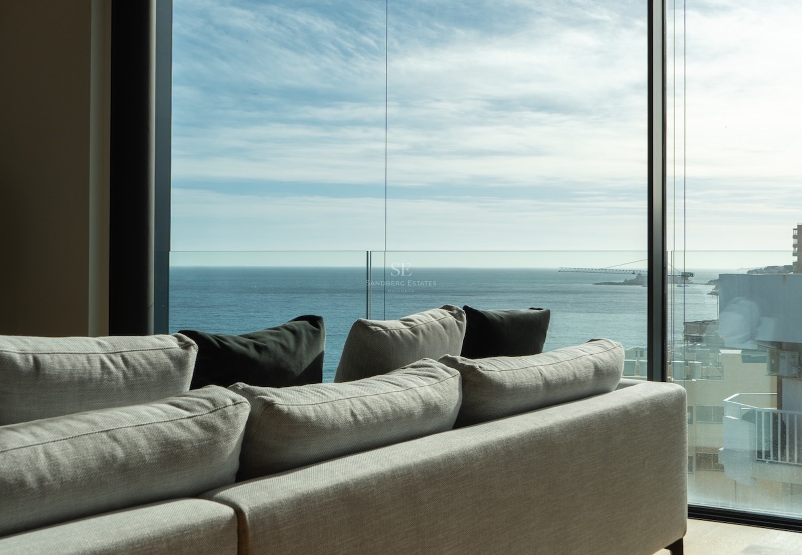 Modernes Sofa vor bodentiefen Glasfronten mit weitem Blick auf das Mittelmeer und blauen Himmel.