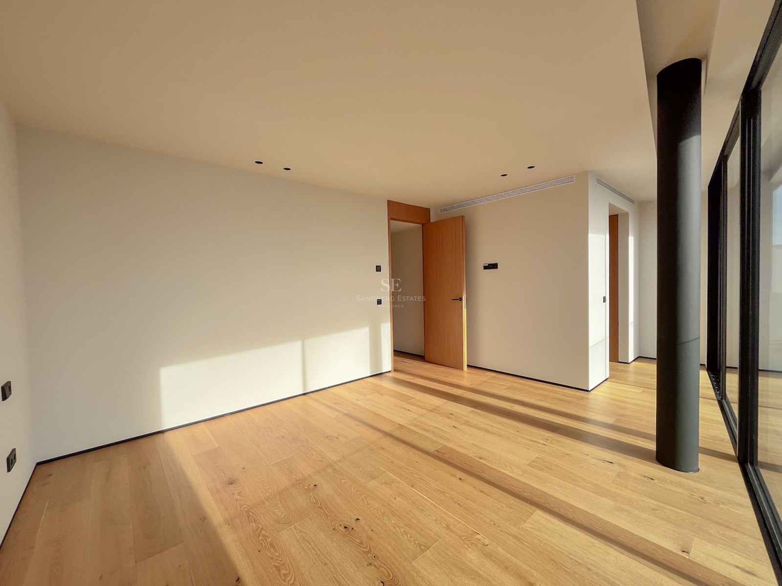 Geräumiges, leeres Schlafzimmer mit hellem Eichenparkett, cremefarbenen Wänden und großen, bodentiefen Glasfenstern.