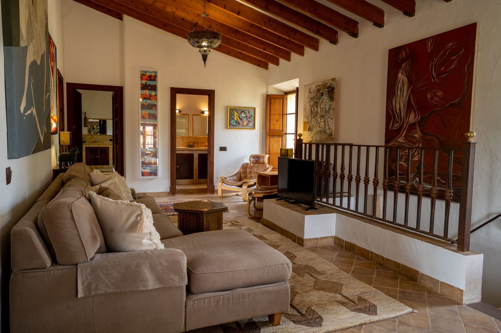 Sala de estar con vigas de madera, suelos de terracota, sofá beige y barandilla de hierro forjado.