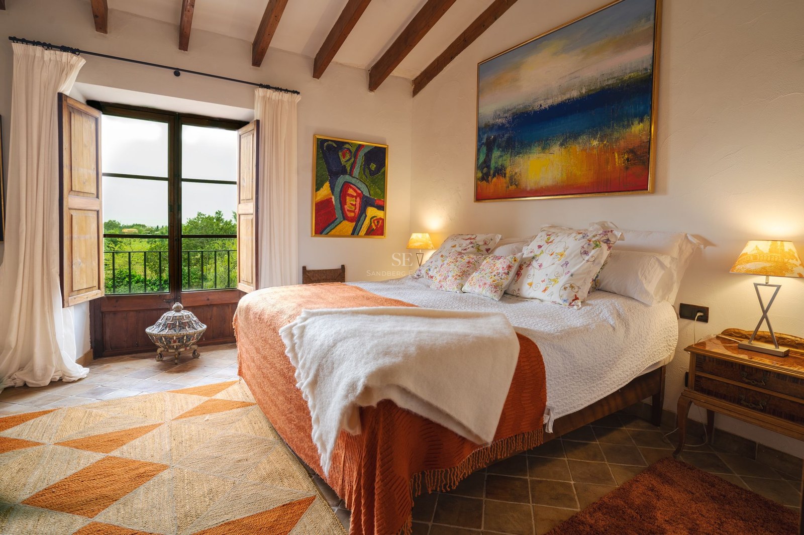 Dormitorio luminoso con vigas de madera expuestas, suelos de terracota y ventanal con vistas a la naturaleza.