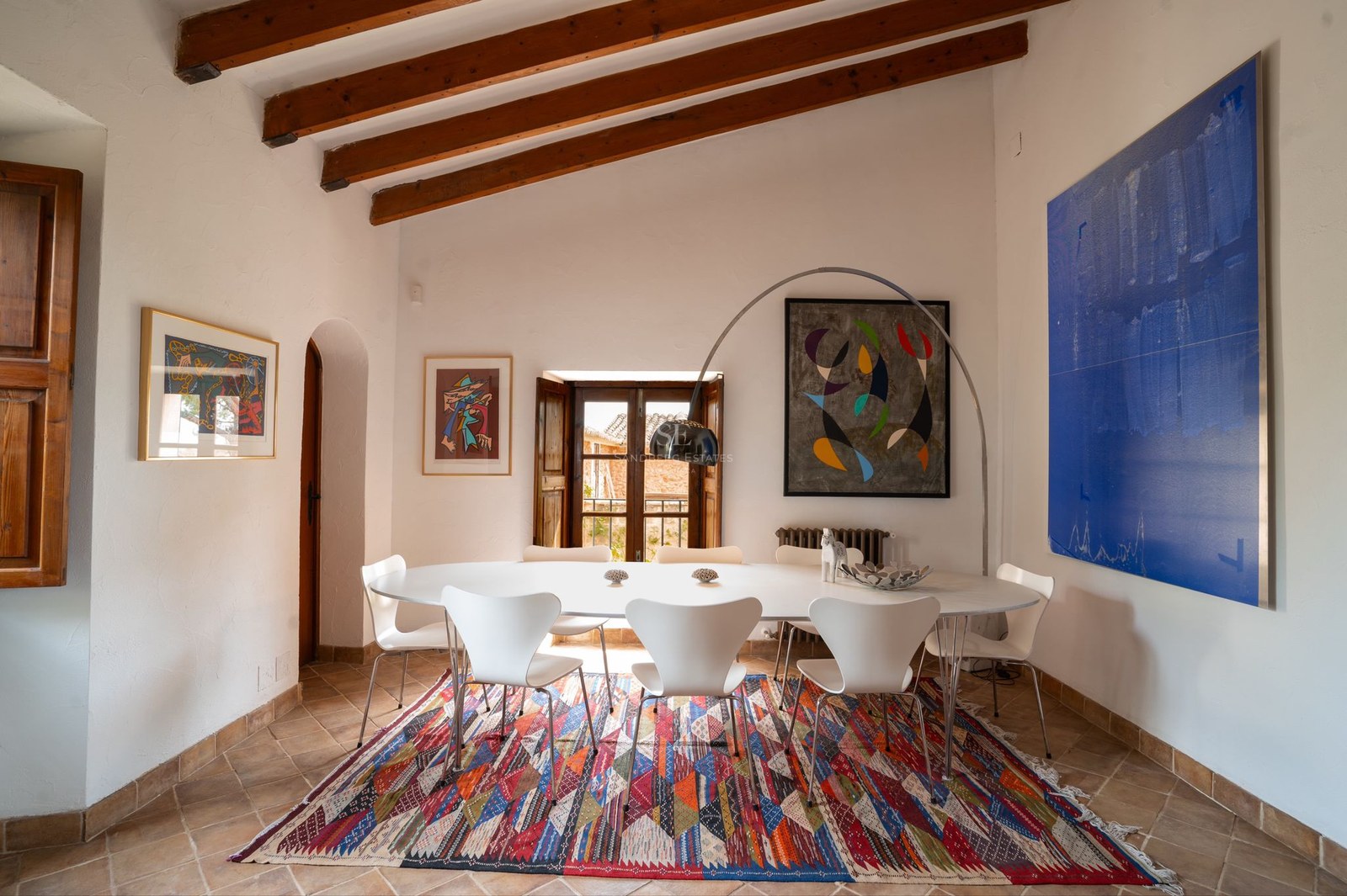 Mesa de comedor blanca con sillas de diseño bajo techos de vigas de madera y suelo de terracota tradicional.