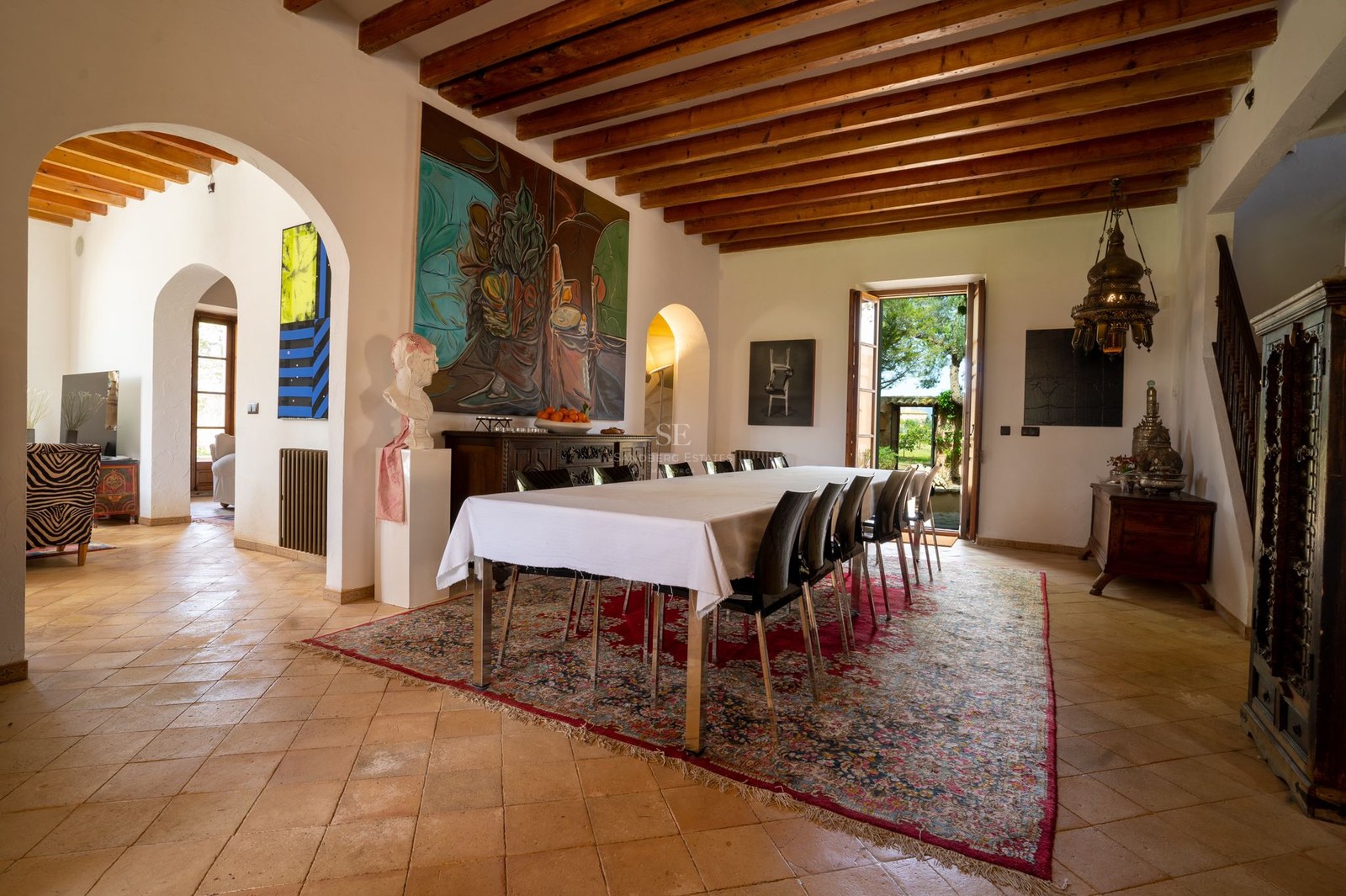Mesa de comedor amplia sobre alfombra clásica en sala con suelos de terracota y vigas de madera.