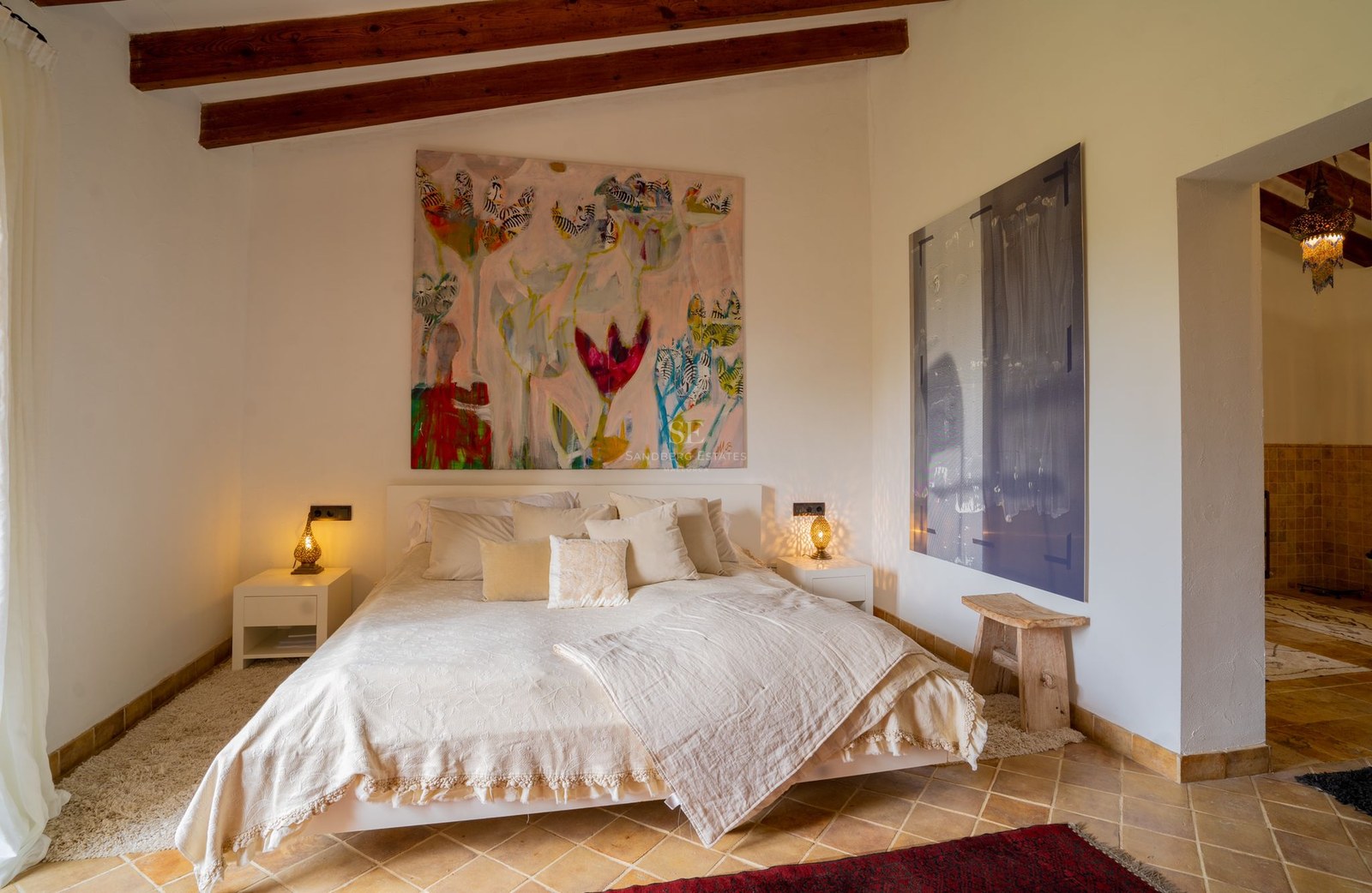 Dormitorio principal con vigas de madera vistas, suelos de terracota, cama amplia y una pintura floral grande.