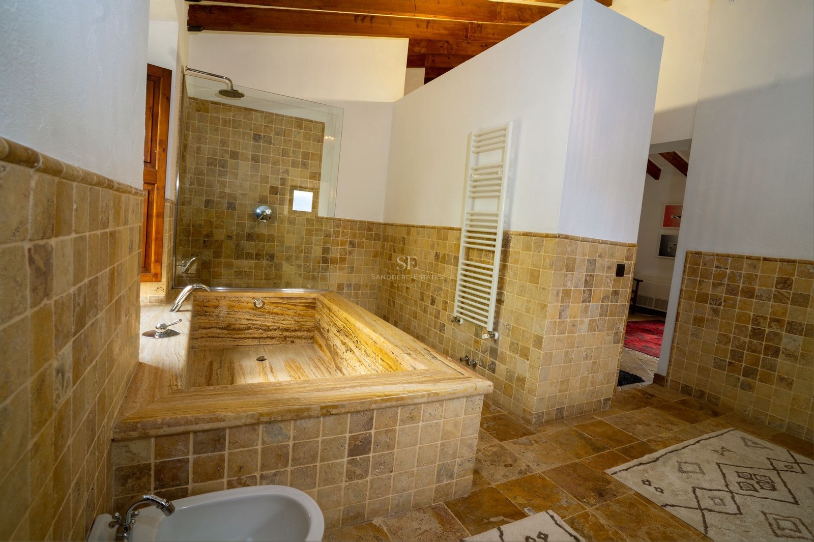 Baño de lujo con bañera integrada de travertino, azulejos de piedra natural y vigas de madera a la vista.