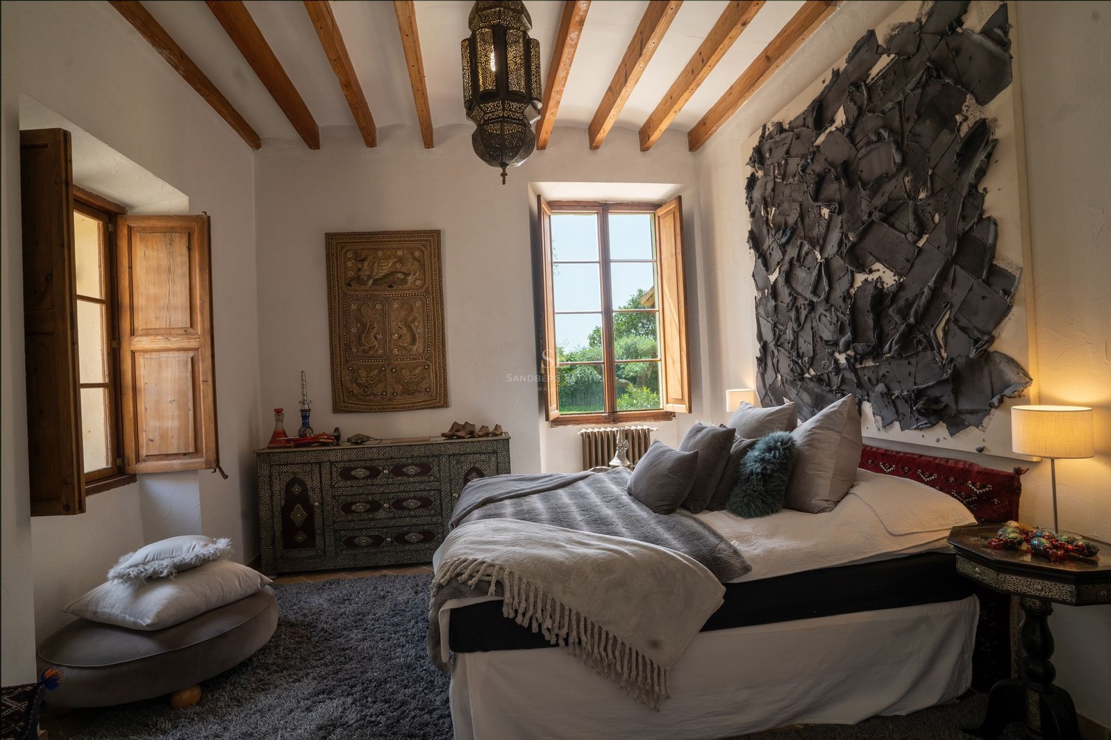 Habitación con vigas de madera vistas, cama amplia con textiles grises y ventana hacia el jardín.