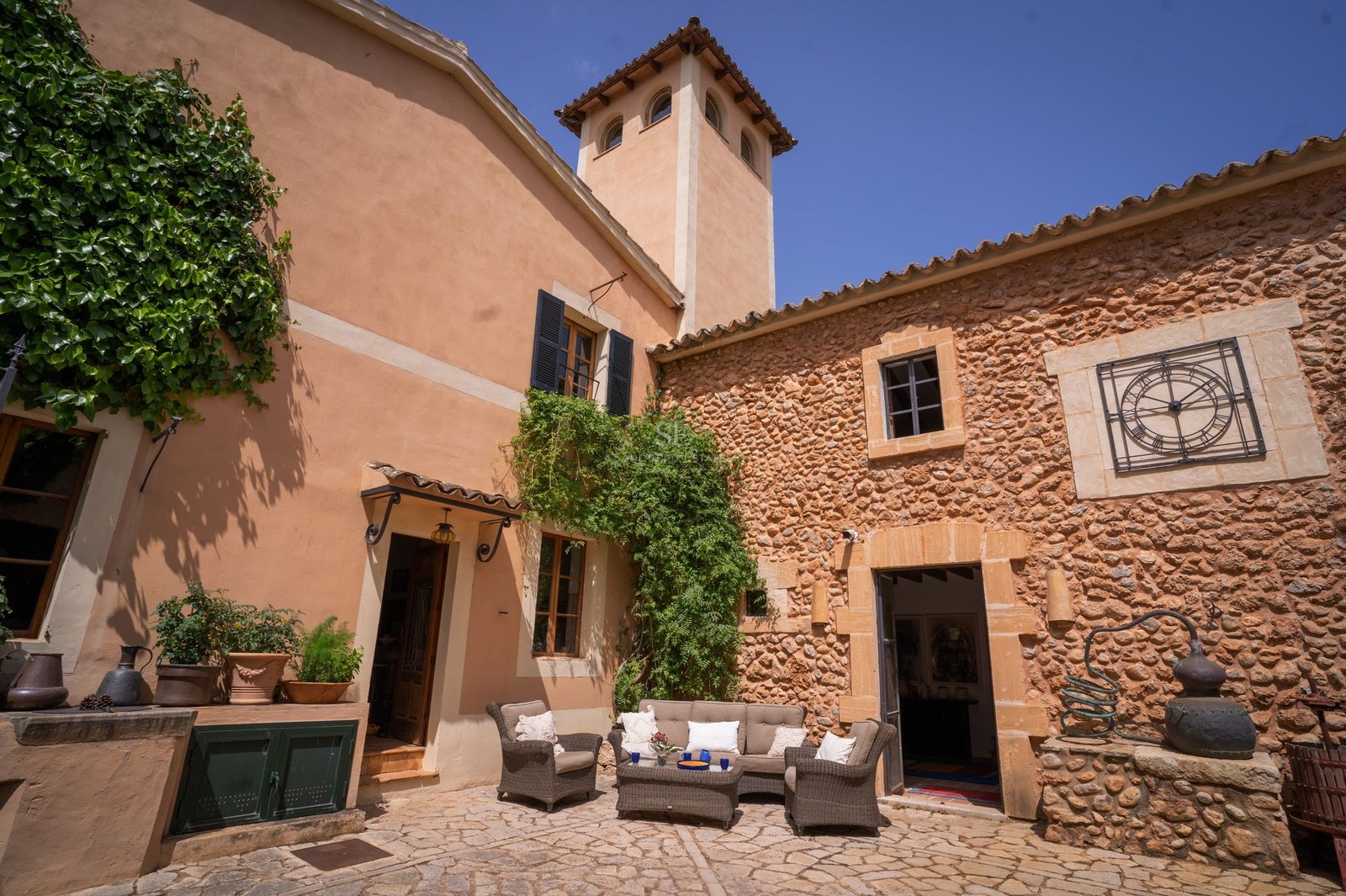 Patio de una villa de piedra con muebles de exterior, paredes de terracota, una torre y hiedra bajo un cielo azul.