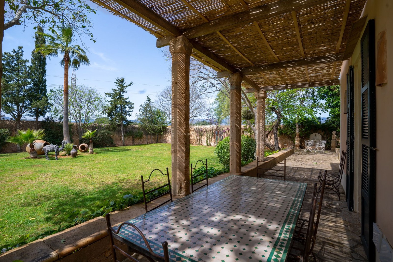 Terraza a la sombra con mesa de mosaico y sillas de hierro frente a un jardín privado con césped y árboles.
