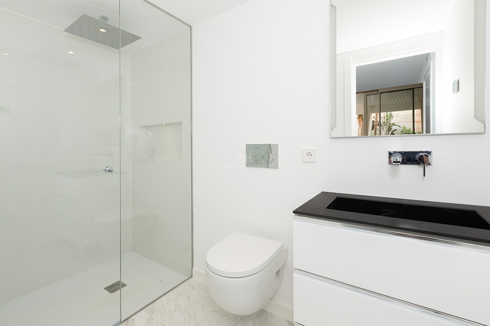 A bright, minimalist bathroom featuring a glass walk-in shower, a white wall-hung toilet, and a black and white vanity.