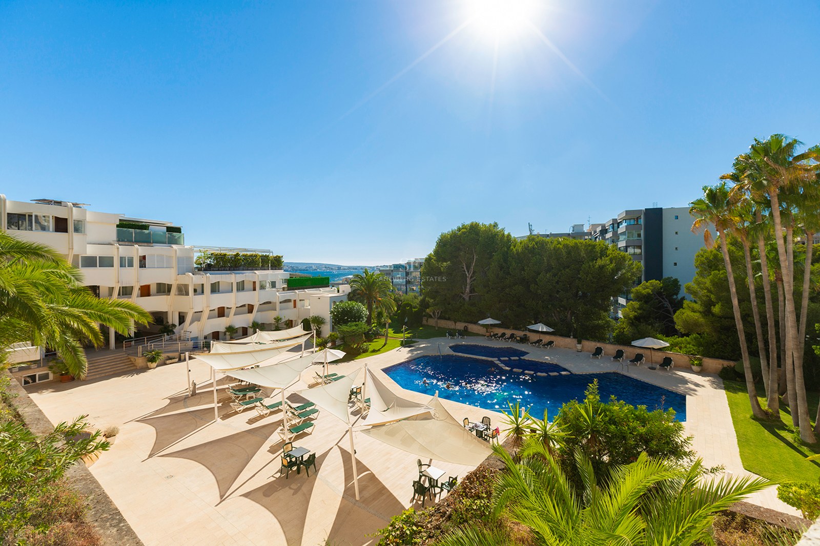 Large blue swimming pool surrounded by beige terrace, white sun sails, palm trees, and resort buildings under a bright sun.
